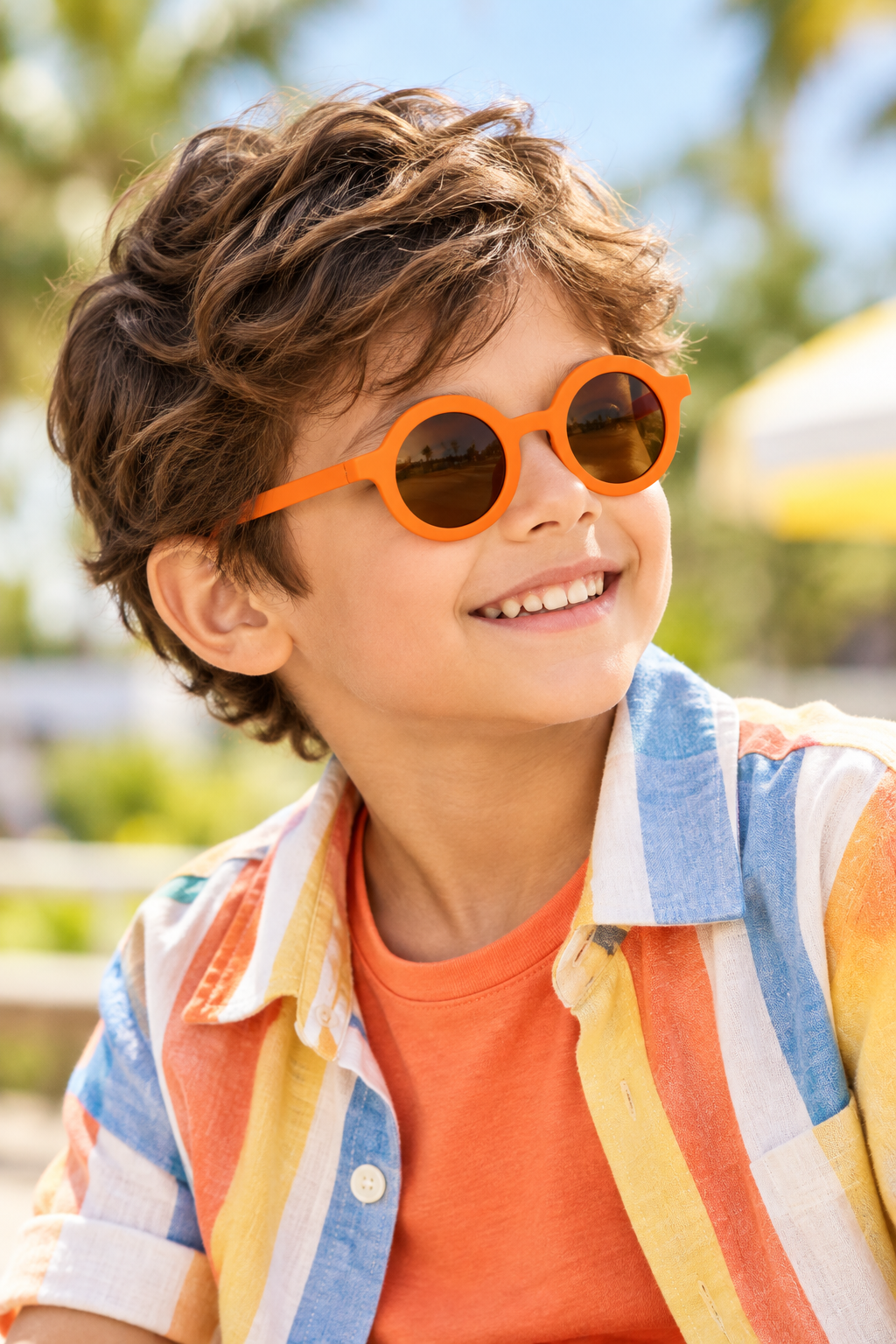 A smiling young boy with wavy brown hair wearing bright orange round sunglasses and a multicolored striped shirt over an orange t-shirt, photographed outdoors with a blurred sunny background.