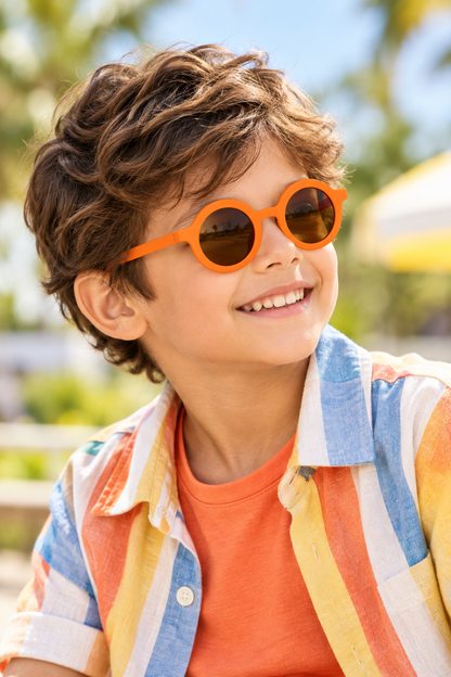A smiling young boy with wavy brown hair wearing bright orange round sunglasses and a multicolored striped shirt over an orange t-shirt, photographed outdoors with a blurred sunny background.