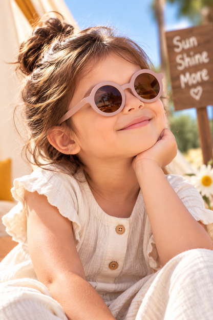 A young girl with her hair in a bun wearing round light pink sunglasses and a cream-colored ruffled top, resting her chin on her hand in a sunny outdoor setting.