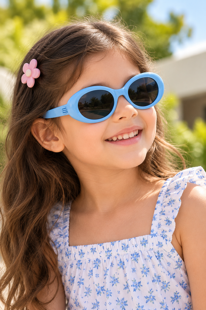 A young girl with long brown hair wearing light blue oval sunglasses and a white floral dress, photographed outdoors with a blurred green background.