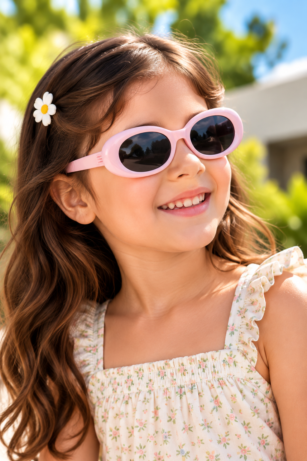A young girl with long brown hair wearing pink oval sunglasses and a white floral top, smiling outdoors with green foliage in the background.
