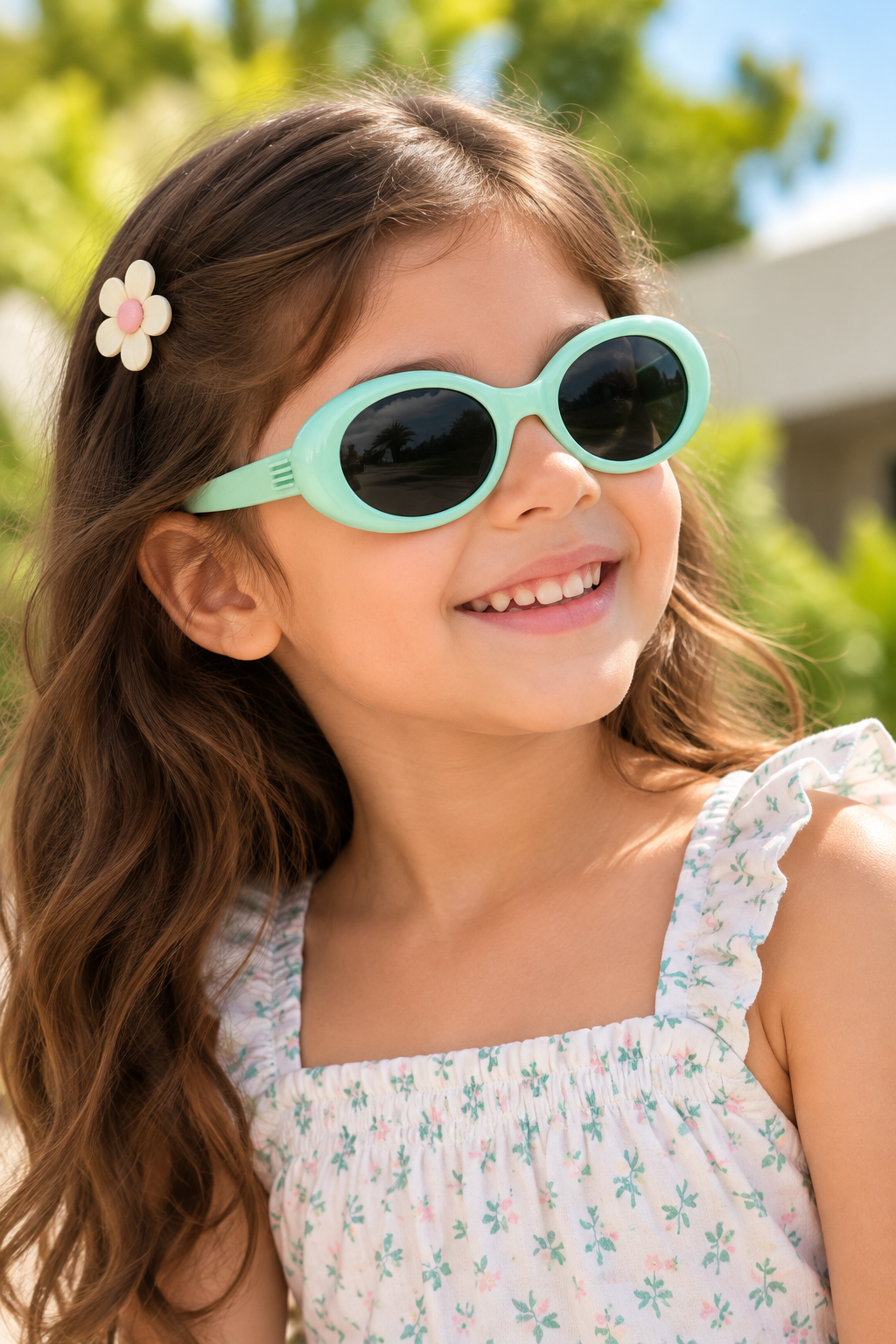 A young girl with long brown hair wearing mint green oval sunglasses and a white floral top, smiling outdoors with a blurred green background.
